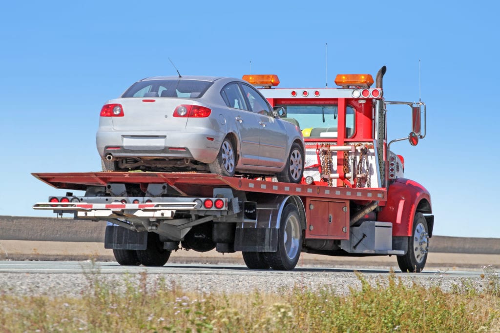 Garage insurance tow truck towing insurance. View or rear quarter of a red tilt bed tow truck on a highway carrying a gray sedan.