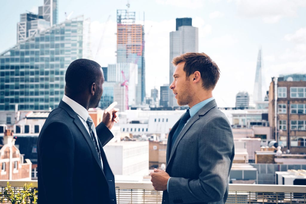 Property Management Insurance landlord client building coverage. Two businessmen in suits overlooking cityscape pointing to managed properties.