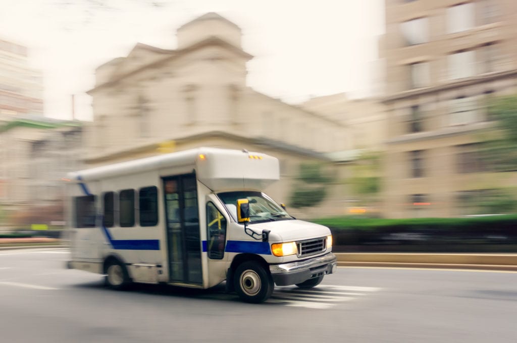 Business auto insurance for assisted living facilities, a small white bus with a blue stripe carries residents on a city street with motion blur background.