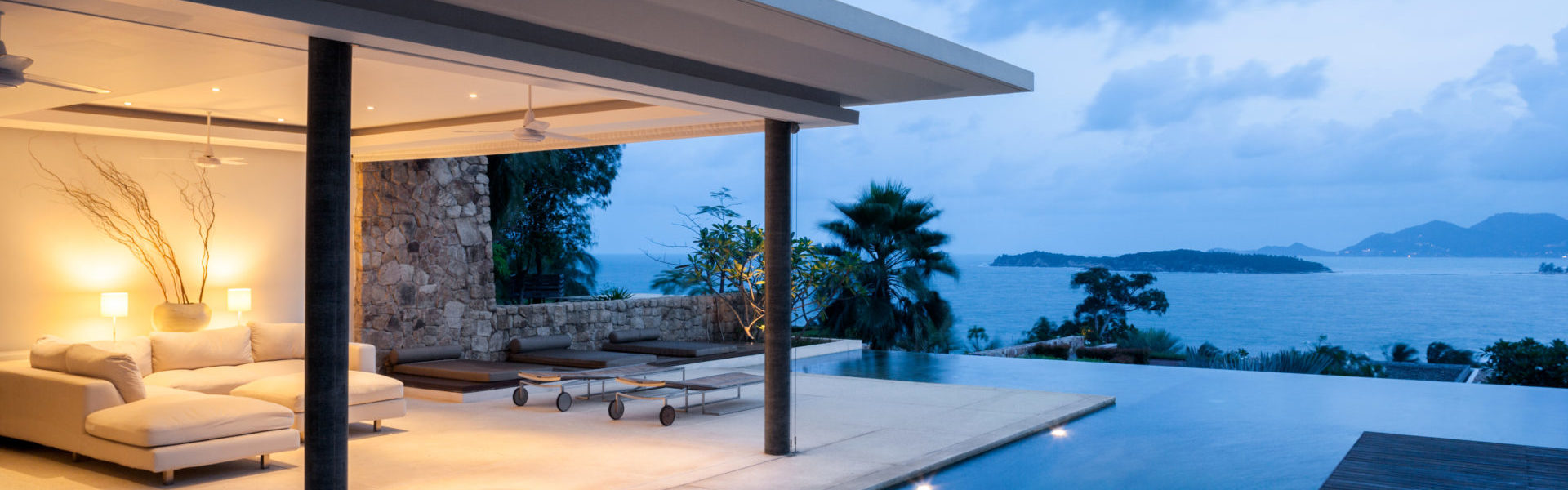 High value home insurance, villa patio scene next to infinity pool at dusk overlooking blue sea with islands in background.