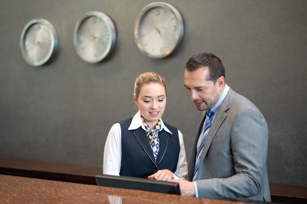 Hotel insurance workers compensation. A male hotel manager in gray suit talks to a woman working at the front desk.