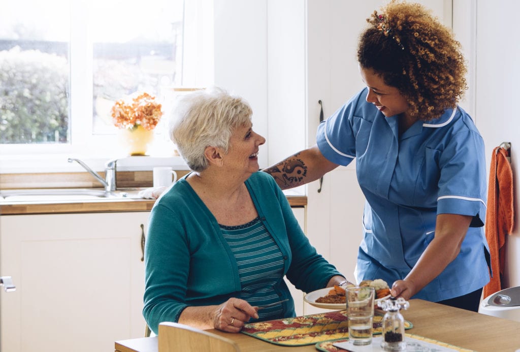 Workers compensation insurance for assisted living facilities, worker helping a woman at kitchen table, serving her a meal.