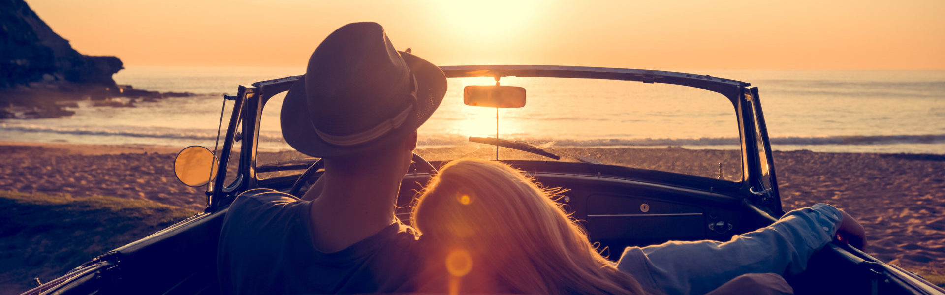 Luxury car insurance, couple watching an orange sunset over a beach in a red convertible car.