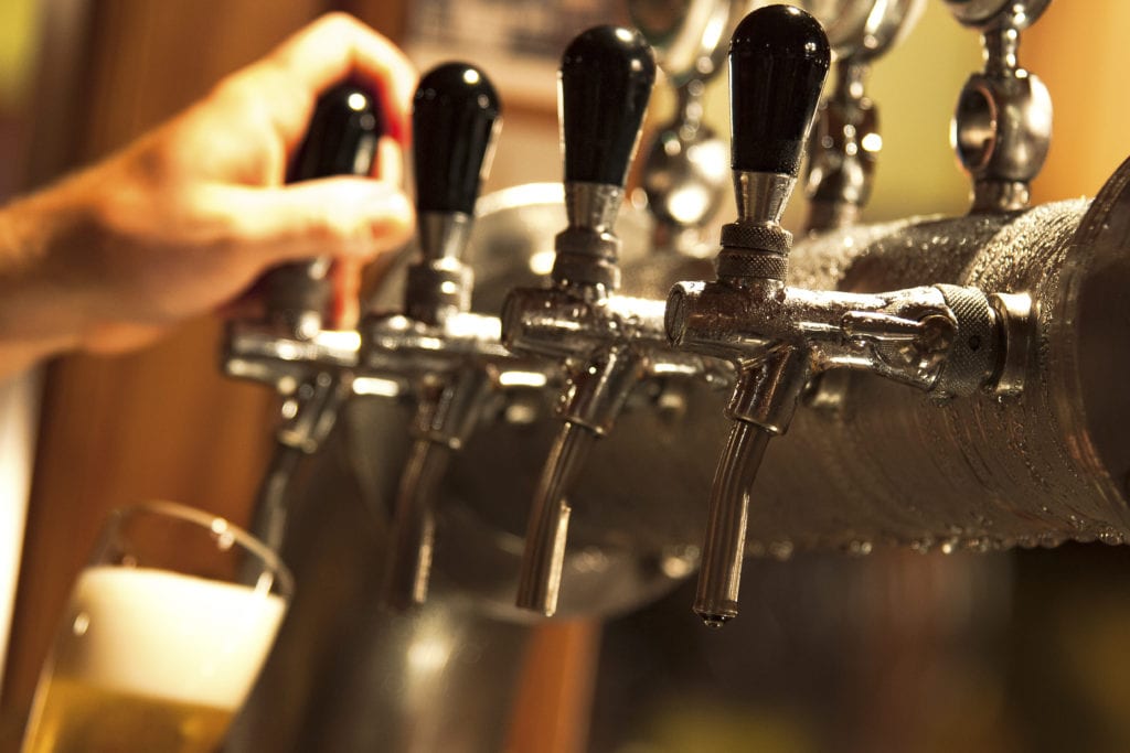 Craft brewery liability insurance, a barman fills a beer glass from a row of stainless steel taps, pipes dripping with condensation.