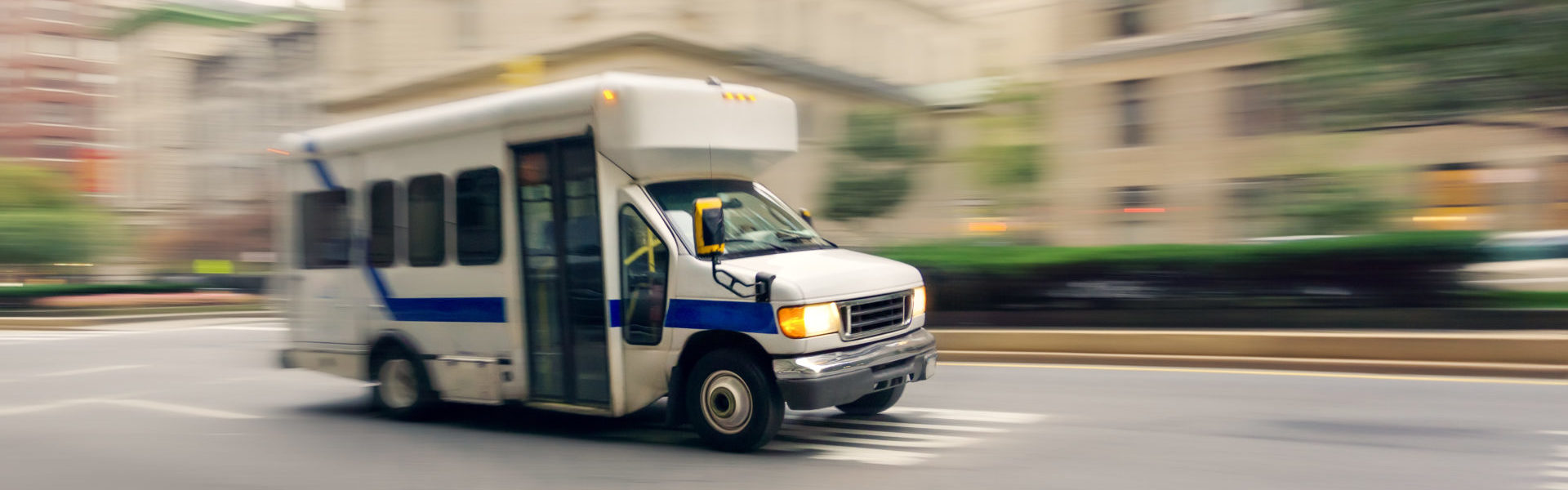 Public transportation insurance NEMT header, a small non-emergency transportation bus on a city street with motion blur background.
