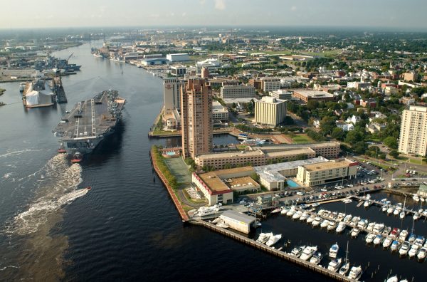 Aerial view of tug boats guiding USS Harry S Truman up the Elizabeth River past marina and tall hotels.