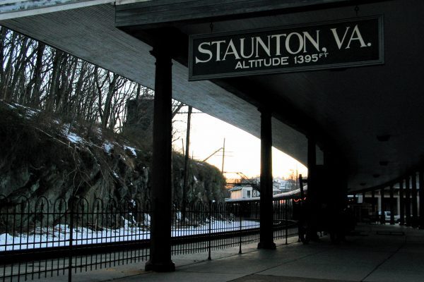 Staunton, VA train station, covered passenger area with sign stating altitude of 1395 feet, next to train tracks, decorative black iron fence separating them.