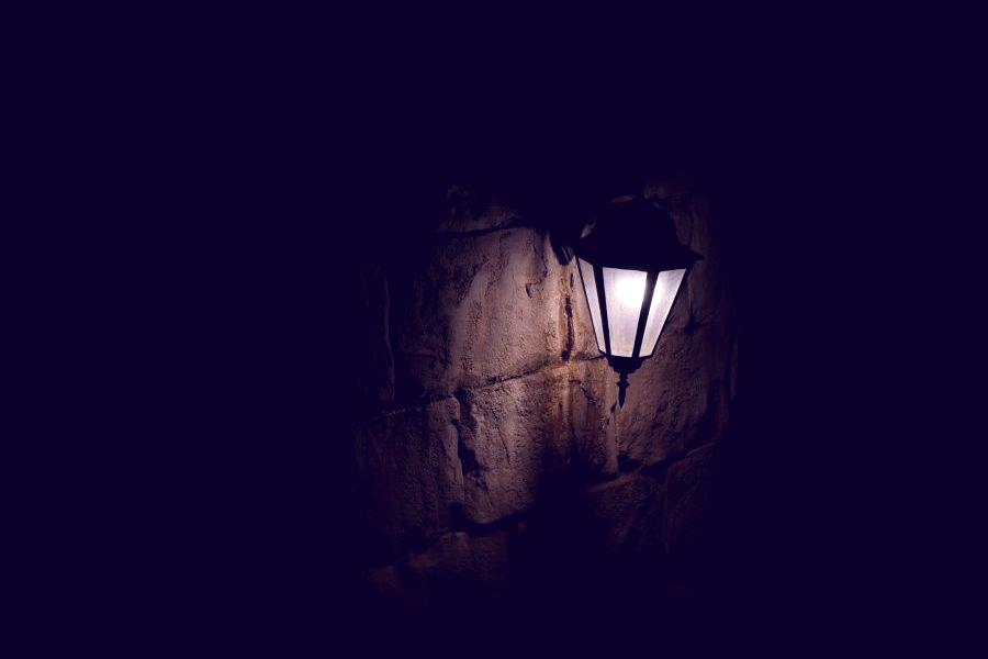 A dark lamp on an exterior stone wall that is flickering and buzzing indicating a wiring issue.
