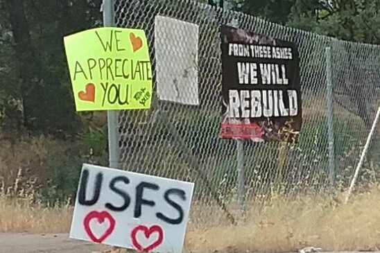 Paper signs on chain link fence announcing We Appreciate You, USFS, and From These Ashes We Will Rebuild.